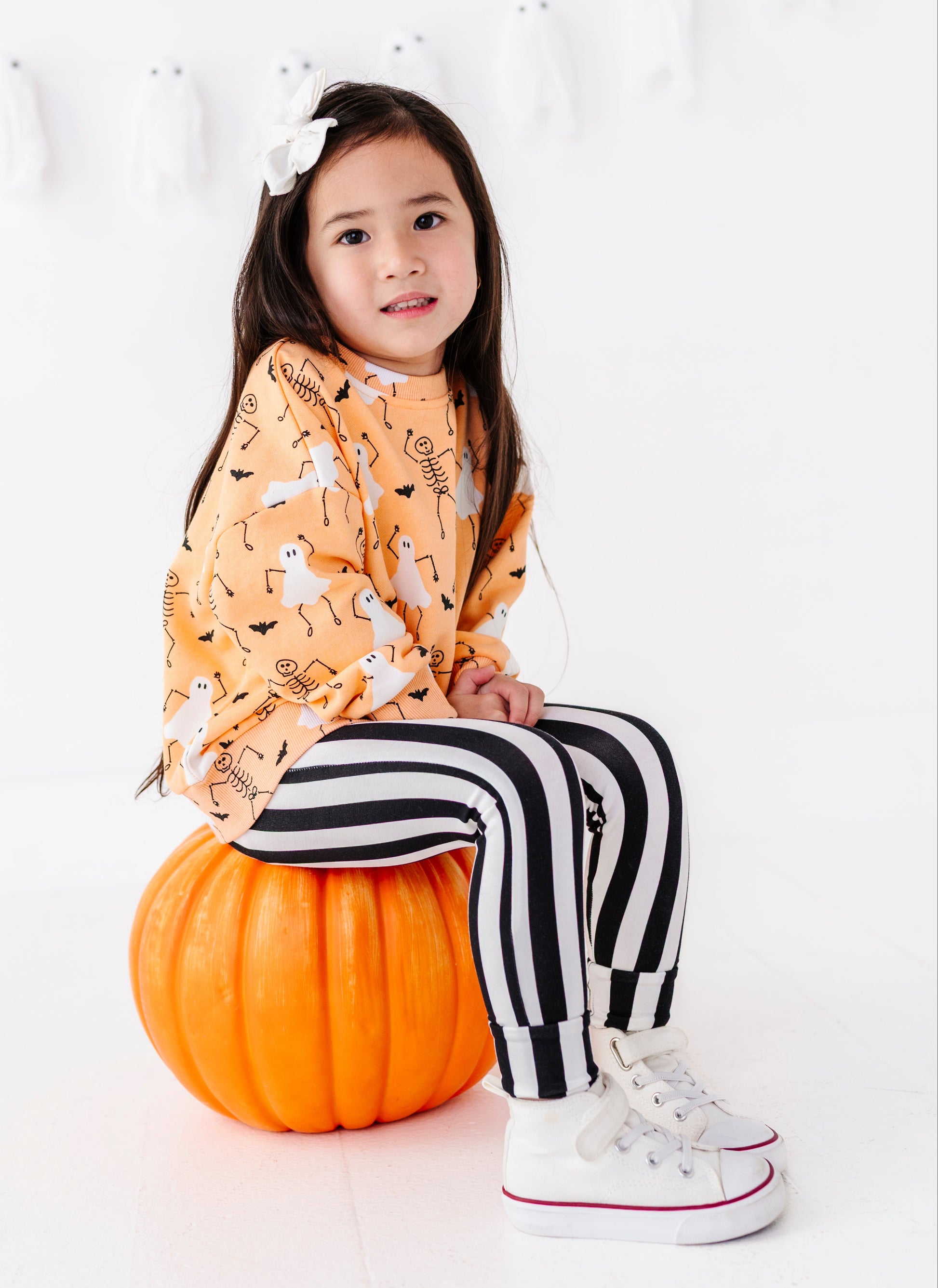 A young girl sits on a large orange pumpkin in a bright white studio setting. She is wearing a long-sleeve orange top with a white ghost and black bat pattern, black and white vertically striped pants, and white sneakers with red trim.