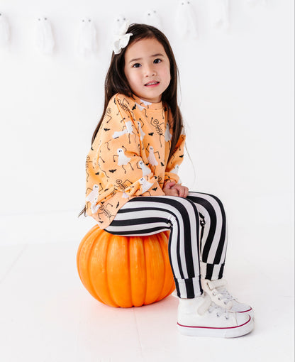 A young girl sits on a large orange pumpkin in a bright white studio setting. She is wearing a long-sleeve orange top with a white ghost and black bat pattern, black and white vertically striped pants, and white sneakers with red trim.