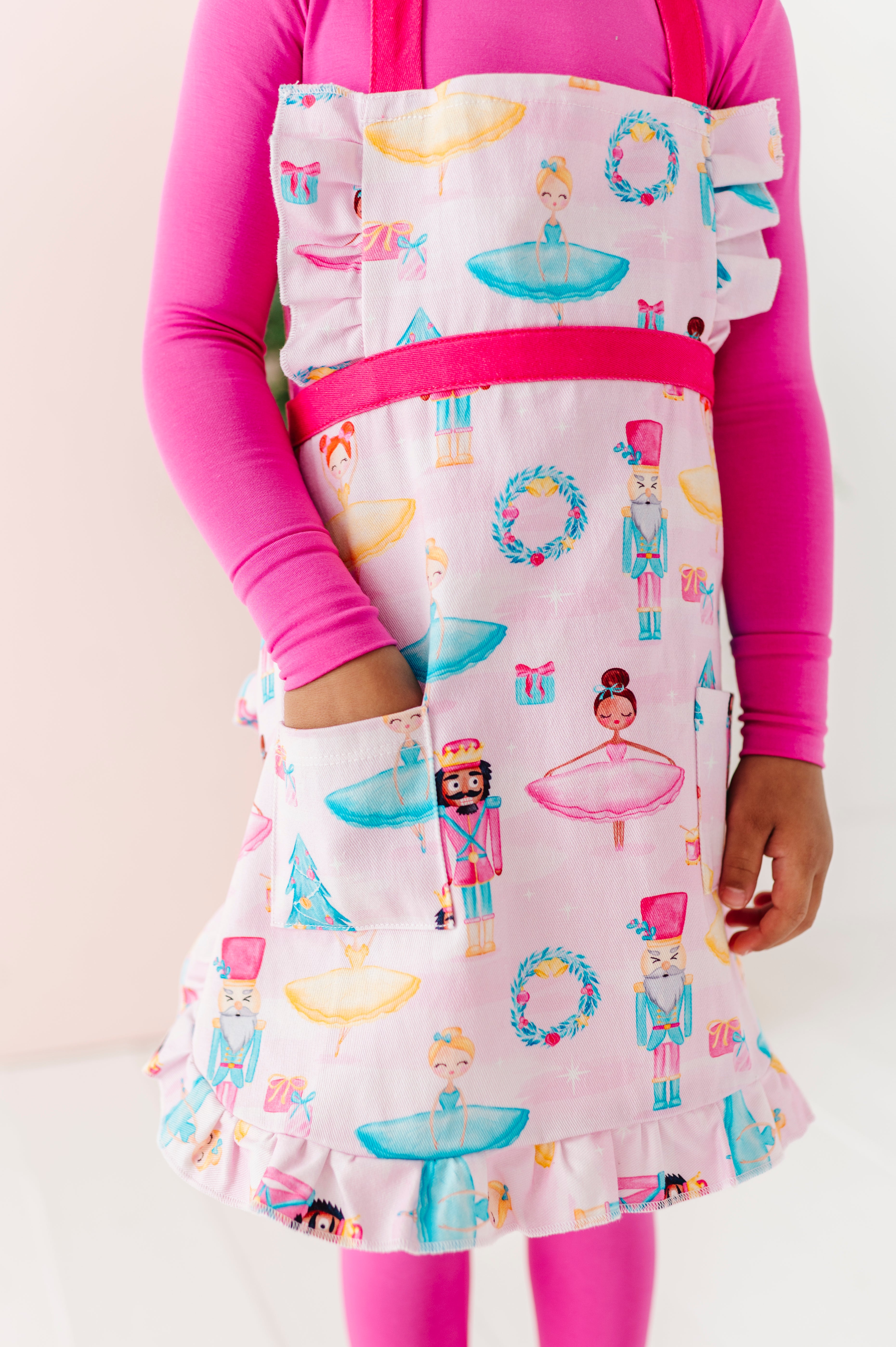 Child wearing a light pink apron featuring ballerinas, nutcrackers, wreaths, presents, and Christmas trees