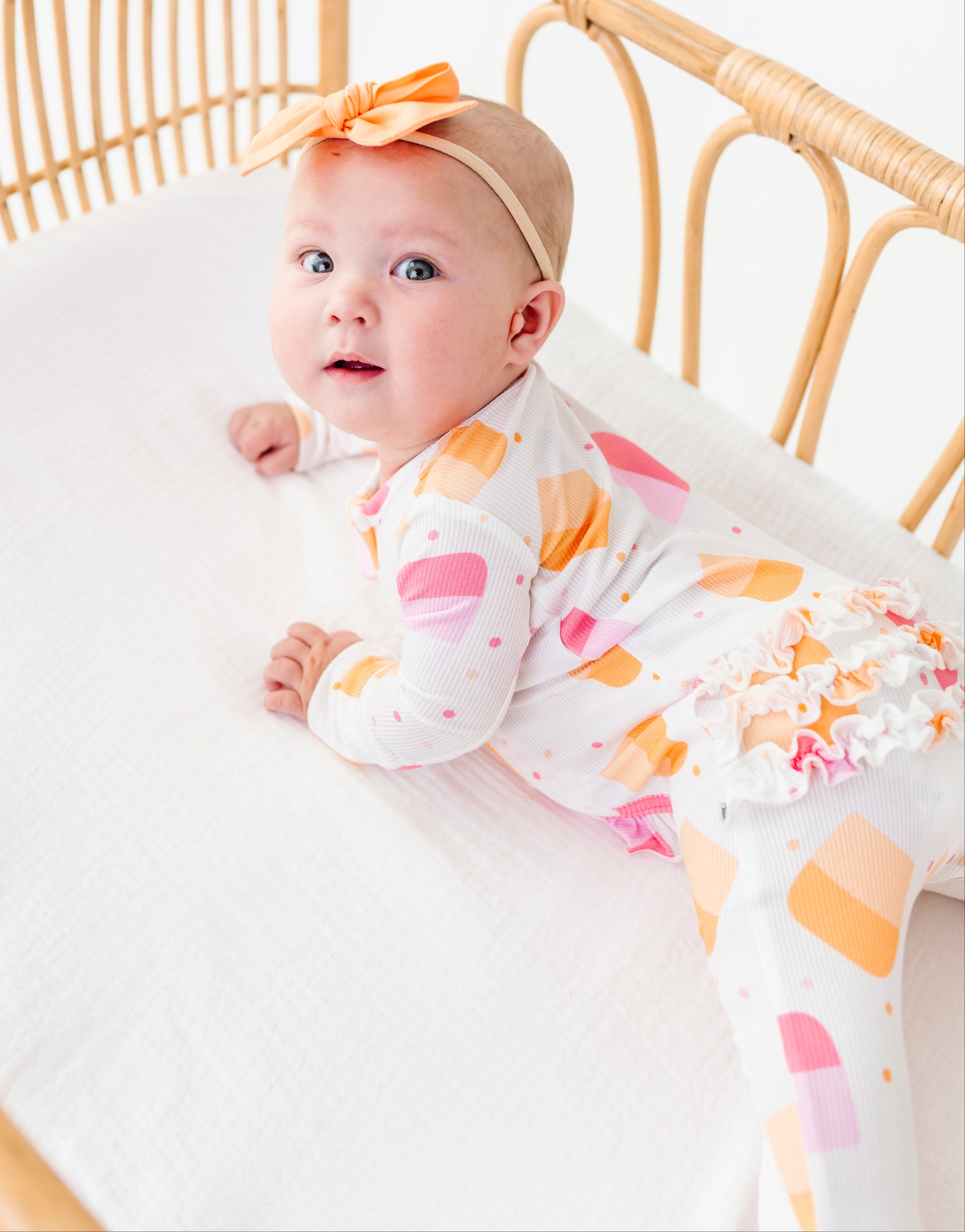 A baby lying on their stomach in a rattan bassinet, wearing a white long-sleeve footed sleeper with a colorful ice cream cone pattern in pink, orange, and yellow, along with an orange bow headband.