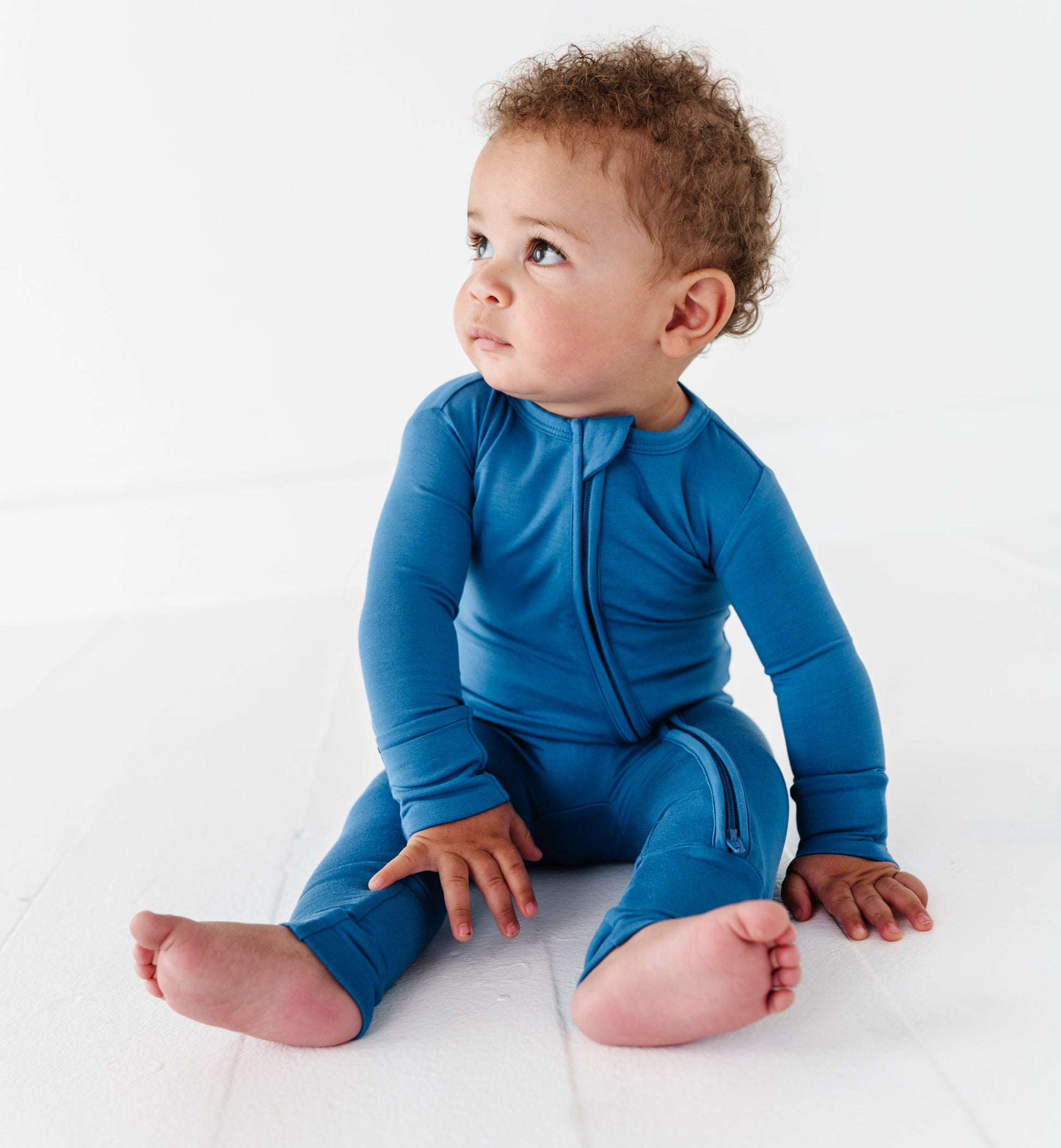Baby wearing a blue bamboo zip romper sitting on a white surface