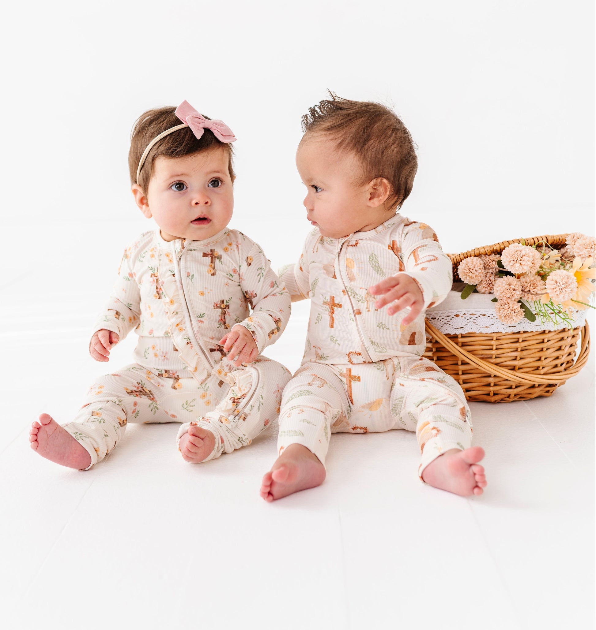 Two babies in matching Easter bamboo pajamas sitting on a white surface with a basket of flowers.