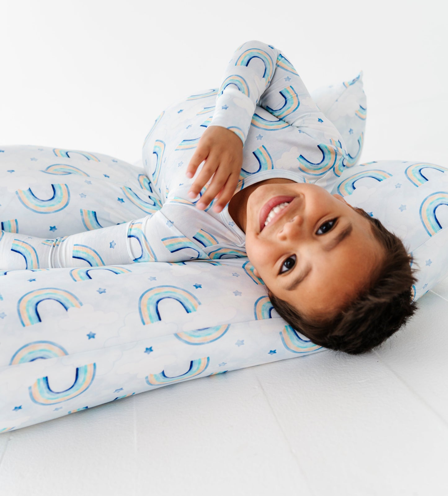 Child wearing a rainbow themed pajama set and resting on a set of matching watercolor rainbow pillowcases on a white background
