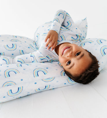 Child wearing a rainbow themed pajama set and resting on a set of matching watercolor rainbow pillowcases on a white background