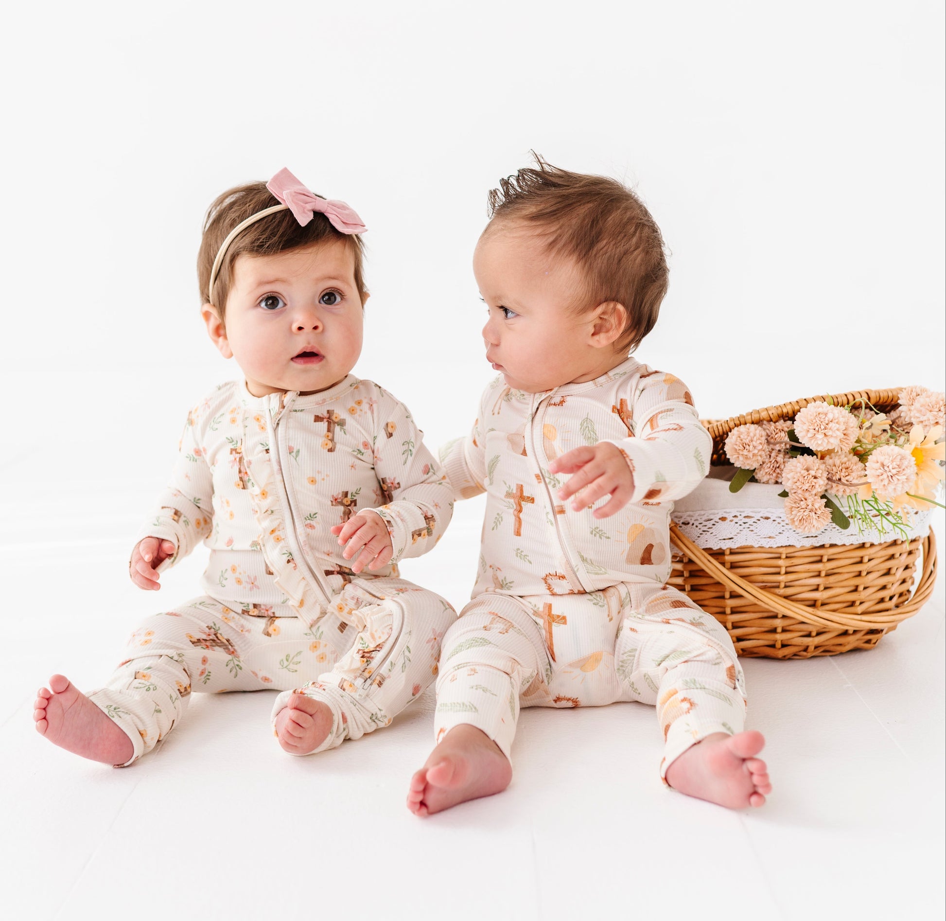 Two babies in matching Easter pajamas sitting on a white surface with a basket of flowers.