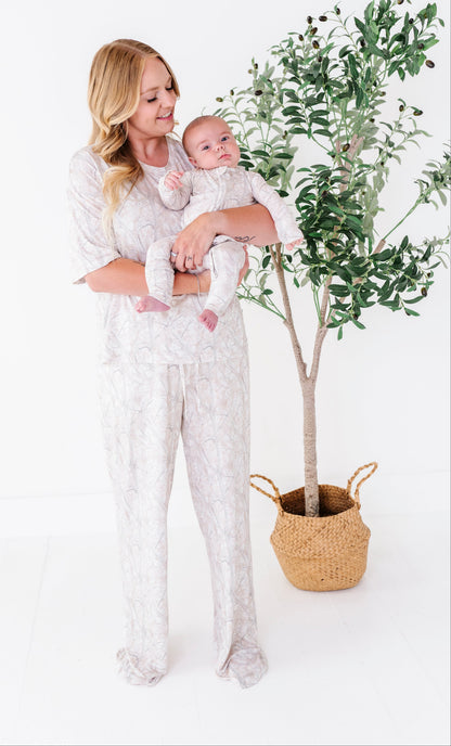 Woman in a camo lounge set holding a baby next to a potted plant on a white background