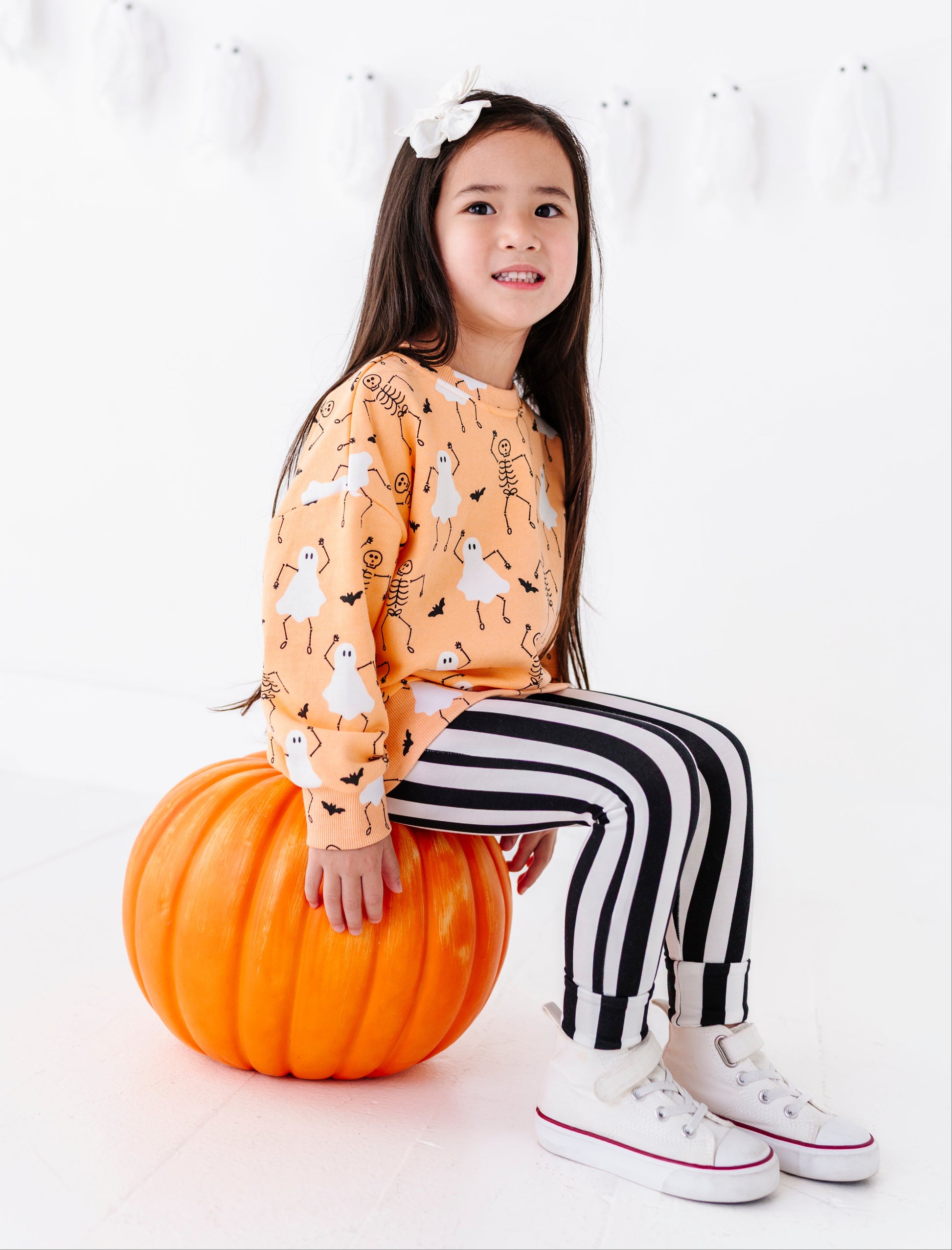 A young girl with a white bow in her hair sits on a large orange pumpkin, wearing a long-sleeve orange sweatshirt with a white cartoon pattern, black and white striped pants, and white sneakers with red trim, against a white background with a ghost garland.