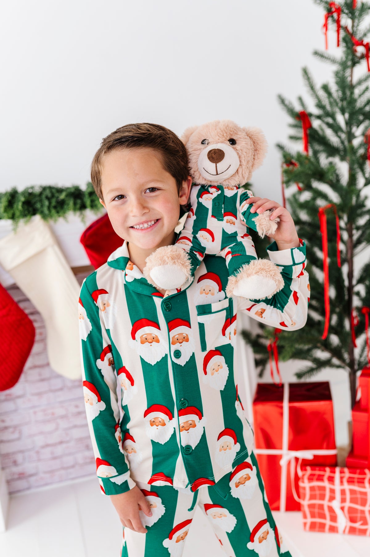 A young boy smiling while wearing a green and white Christmas bamboo pajama set with a Santa Claus face print, holding a teddy bear in matching pajamas, with a decorated Christmas tree and stockings in the background.
