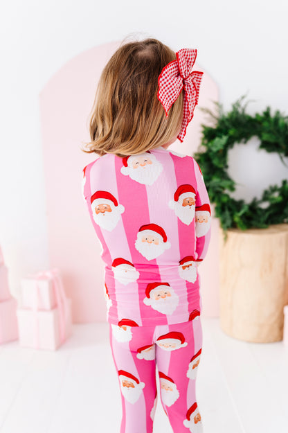A child viewed from behind wearing a pink Santa Claus patterned bamboo pajama set, featuring a red and white gingham bow in their hair, set against a soft pink and white holiday background.