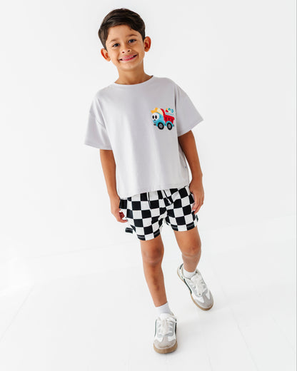 A young boy modeling a white t-shirt with a small colorful cartoon truck graphic on the chest, paired with black and white checkered shorts and white sneakers, set against a seamless white studio background.