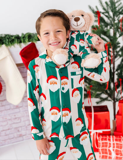 A smiling young boy wearing festive green and white Santa Claus patterned pajamas holds a plush teddy bear dressed in the same holiday pajamas. The background features a decorated Christmas tree, stockings, and wrapped gifts.