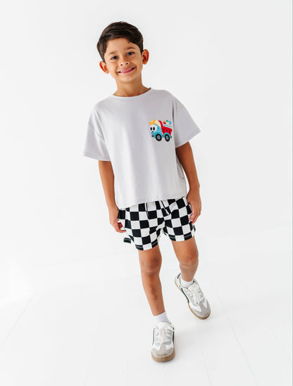 A young boy modeling a white t-shirt with a small colorful cartoon truck graphic on the chest, paired with black and white checkered shorts and white sneakers, set against a seamless white studio background.