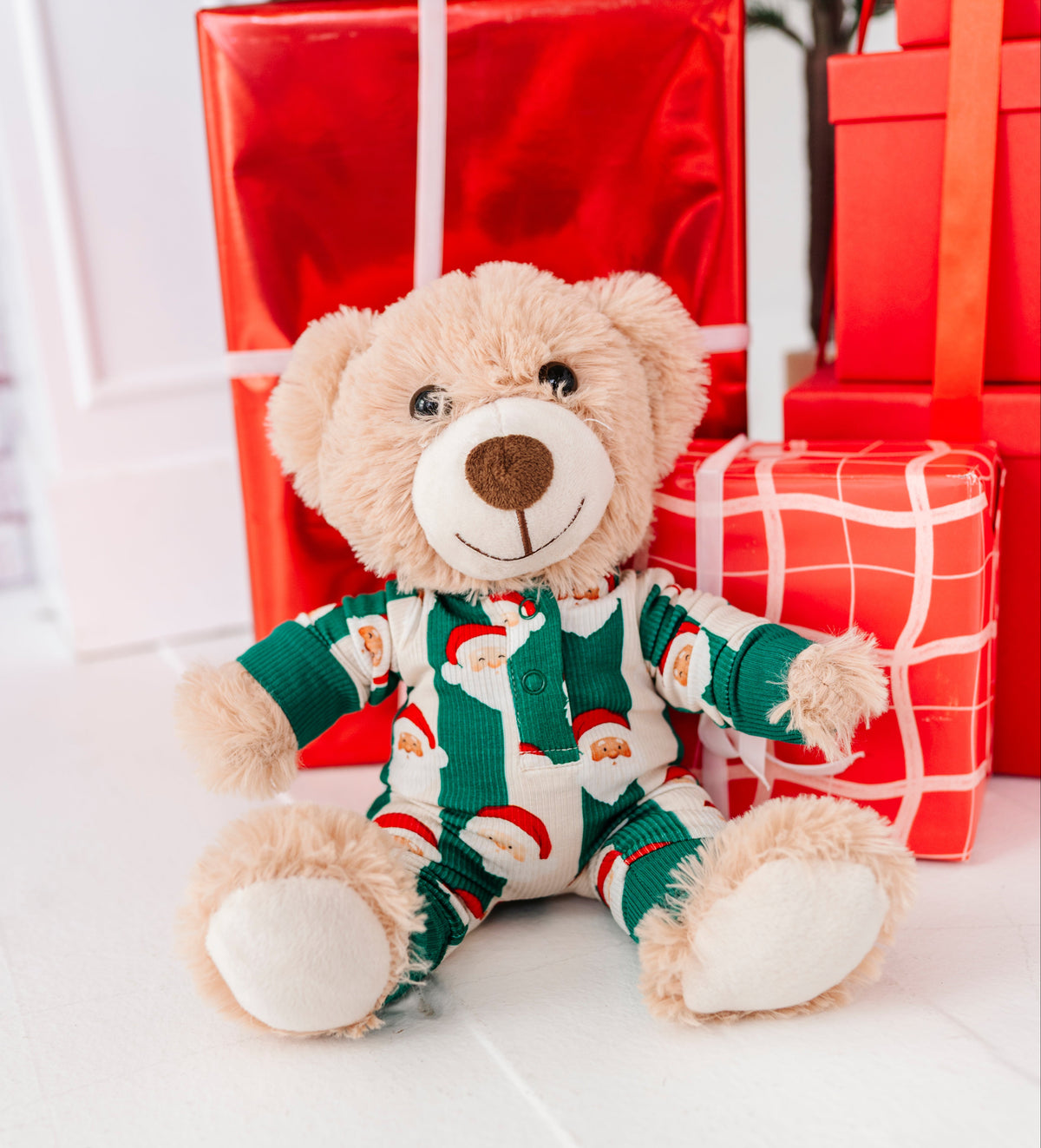 A plush teddy bear wearing green and white Santa-patterned pajamas, seated in front of wrapped Christmas presents on a white floor.