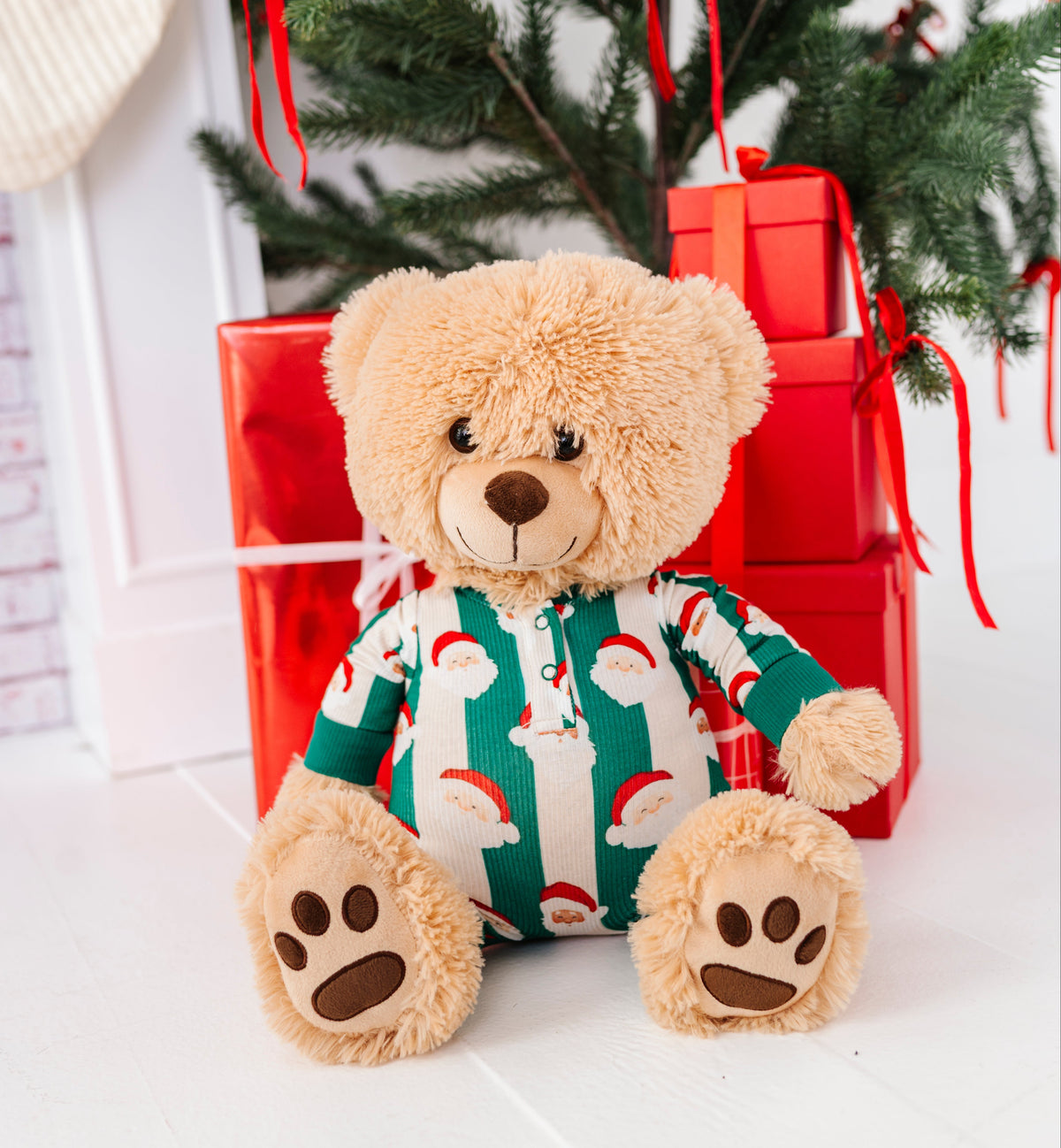 A plush teddy bear sits in front of a Christmas tree and wrapped presents, wearing a green and white Santa-patterned onesie. The scene is styled with holiday decorations on a white floor.