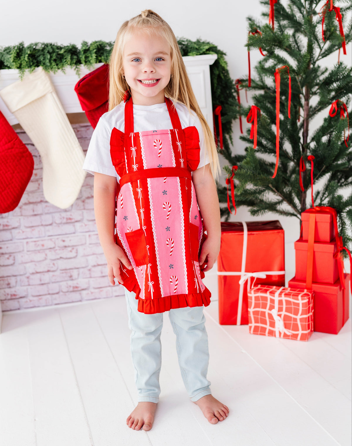 A young girl stands barefoot in a festive holiday setting, wearing a red and pink apron with a candy cane print over a white t-shirt and light blue jeans. The background features a decorated Christmas tree, stockings, and wrapped gifts.