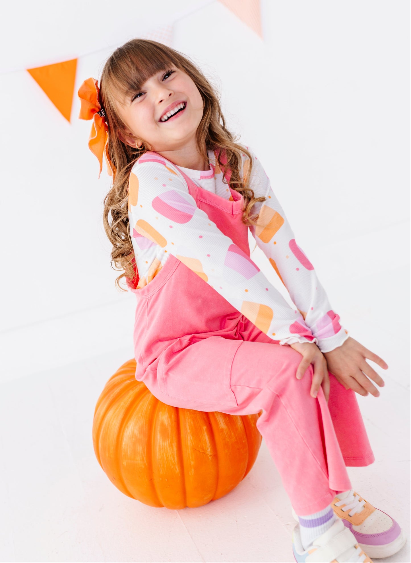 A young girl with a brown hair bow sits smiling on an orange pumpkin, wearing a pink and white long-sleeve outfit with colorful ice cream patterns. The background is a clean white studio setting with orange and pink bunting.