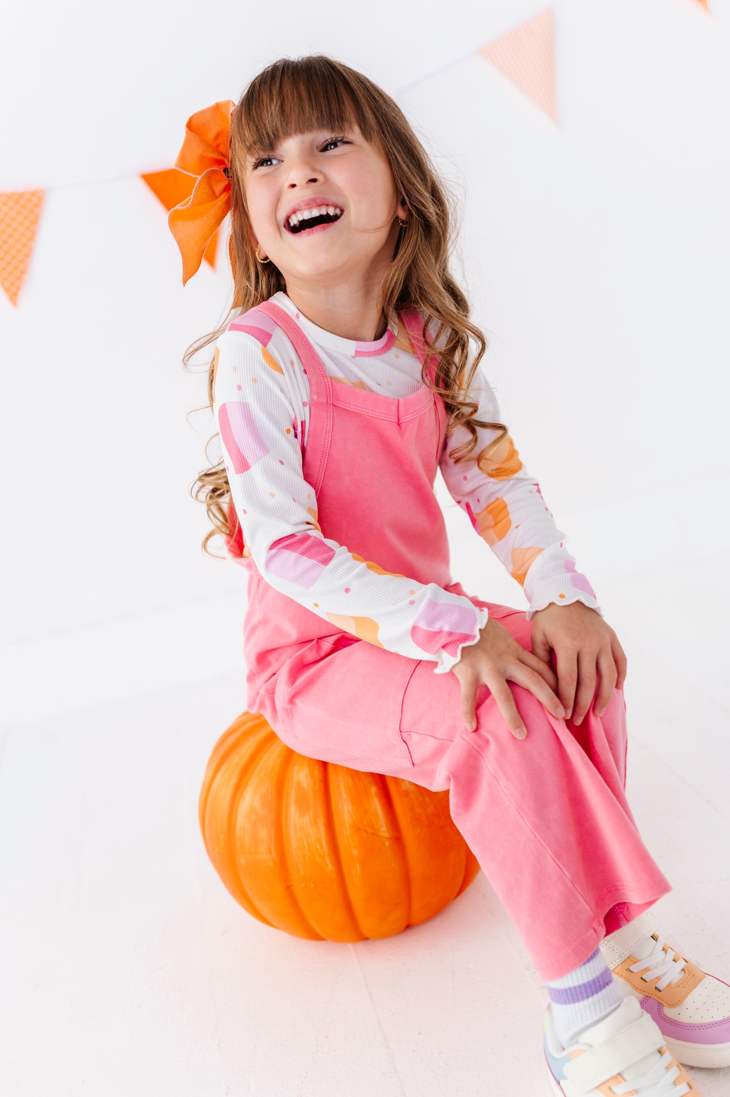 A young girl with long wavy brown hair sits on a large orange pumpkin, wearing a pink overall-style jumpsuit over a white long-sleeve shirt with orange and yellow patterns. She is smiling broadly, set against a bright white background with a decorative orange bow and orange-and-white bunting.