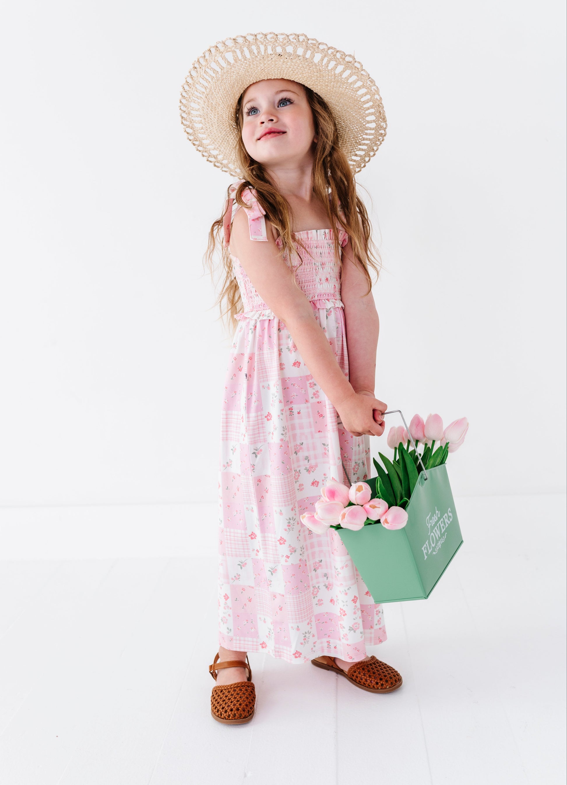Young girl in a pink bamboo maxi dress and straw hat holding tulips against a white background