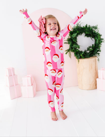 A young girl stands barefoot with her arms raised in the air, wearing a pink Santa Claus patterned two-piece bamboo pajama and a red-and-white bow in her hair. The background is a bright white room decorated with a pink arch, a green wreath, and pink gift boxes.