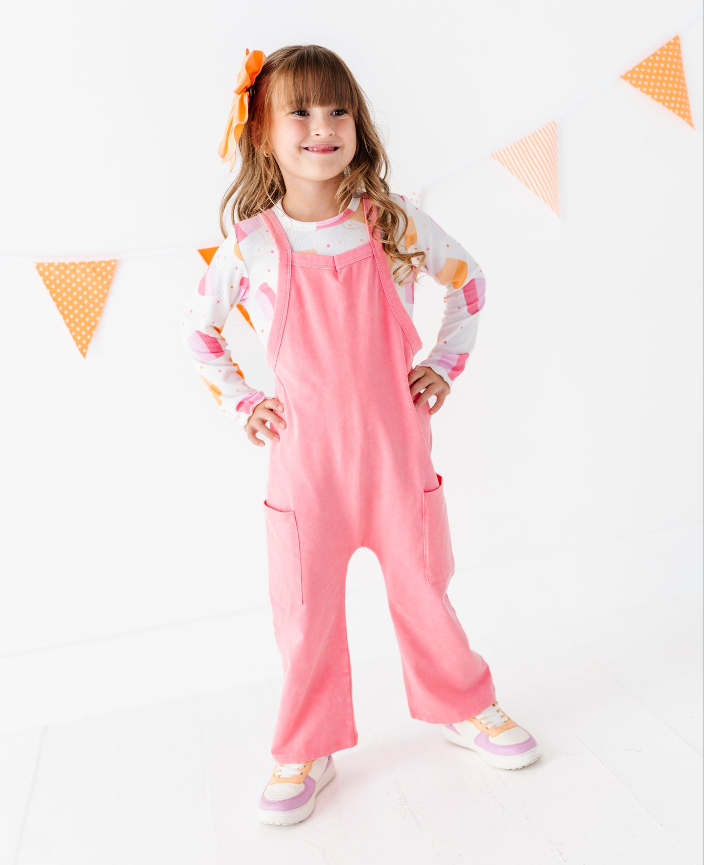 A young girl with a brown hair bow stands in a studio wearing pink overall over a white long-sleeve shirt with colorful graphics. She is posing with hands on hips against a white background with orange and white bunting.