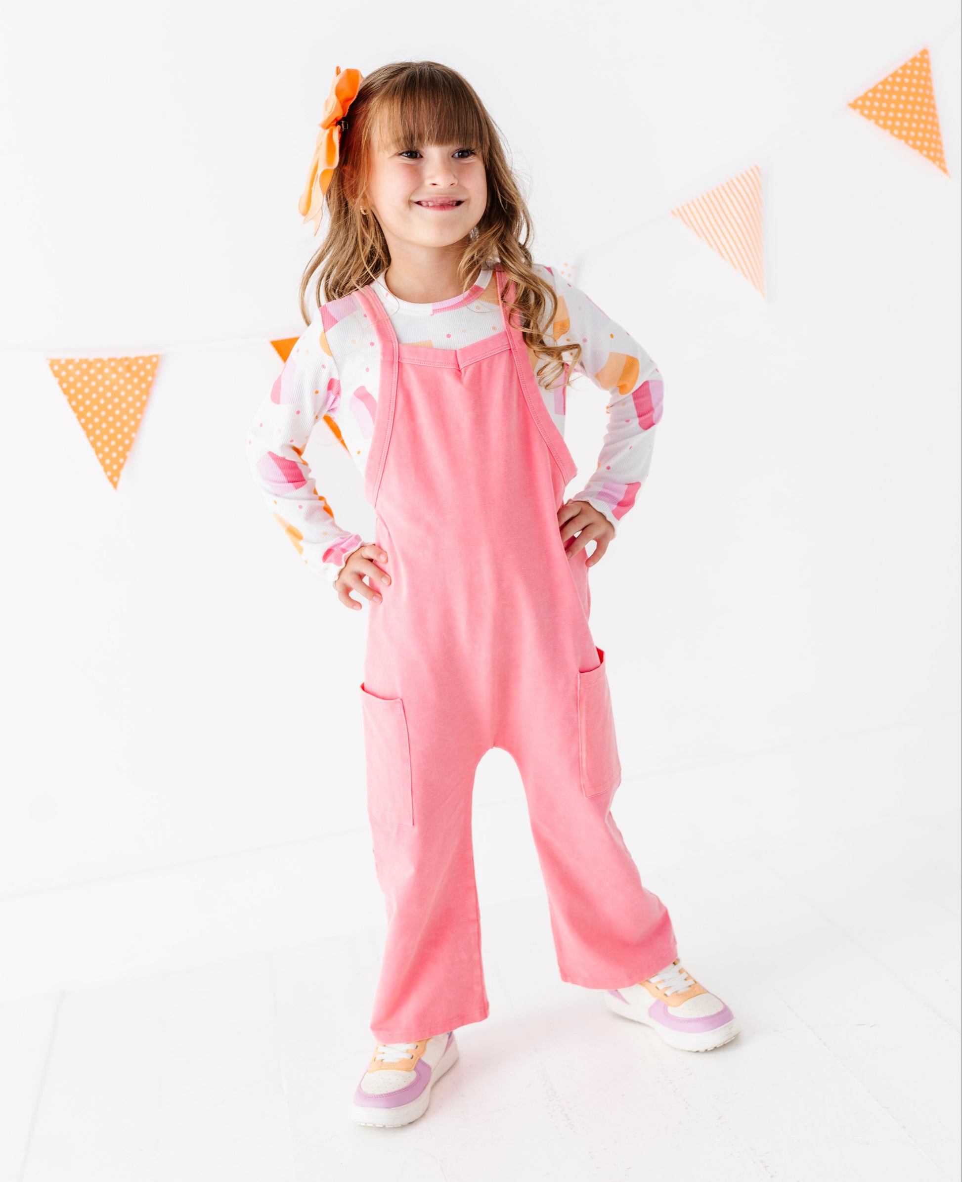 A young girl with a brown hair bow stands in a studio wearing pink overall over a white long-sleeve shirt with colorful graphics. She is posing with hands on hips against a white background with orange and white bunting.