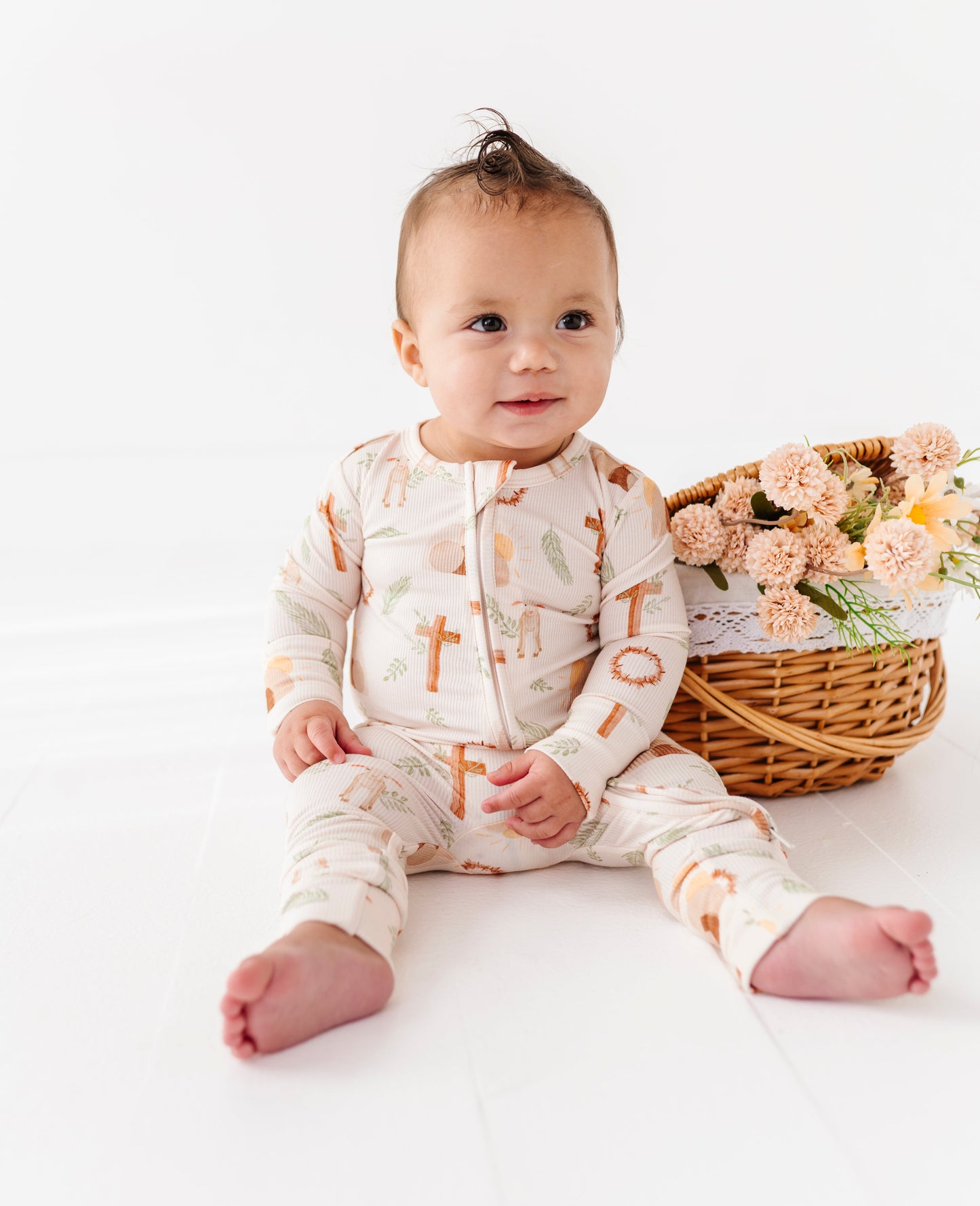 Baby wearing a onesie with printed Easter design, sitting on a white surface with a basket of flowers in the background.
