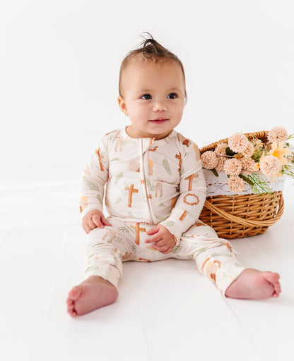 Baby wearing a onesie with printed Easter design, sitting on a white surface with a basket of flowers in the background.
