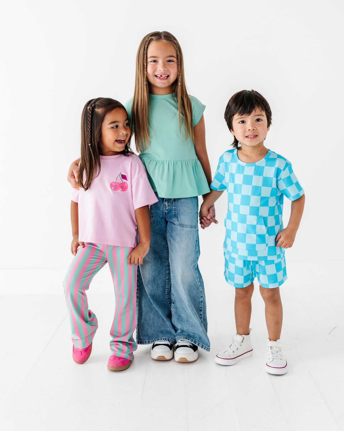 Three children modeling matching casual clothing sets against a clean white studio background. The child on the left wears a pink graphic T-shirt and pink striped pants, the center child wears a mint green peplum top and blue jeans, and the child on the right wears a light blue and white checkered short-sleeve top and matching shorts.