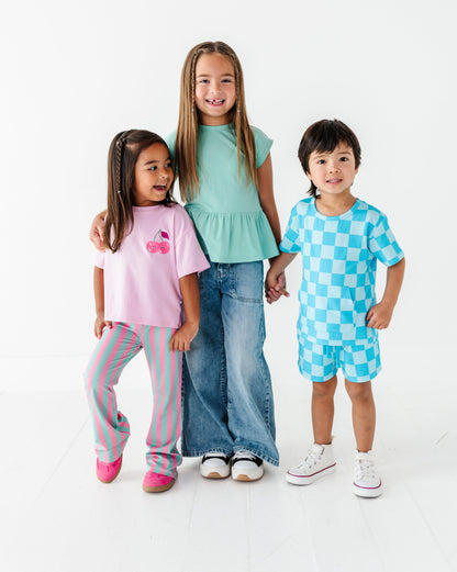 Three children modeling matching casual clothing sets against a clean white studio background. The child on the left wears a pink graphic T-shirt and pink striped pants, the center child wears a mint green peplum top and blue jeans, and the child on the right wears a light blue and white checkered short-sleeve top and matching shorts.