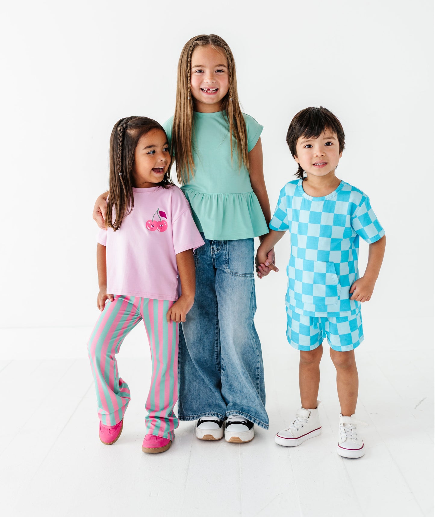 Three children modeling matching casual clothing sets against a clean white studio background. The child on the left wears a pink graphic T-shirt and pink striped pants, the center child wears a mint green peplum top and blue jeans, and the child on the right wears a light blue and white checkered short-sleeve top and matching shorts.