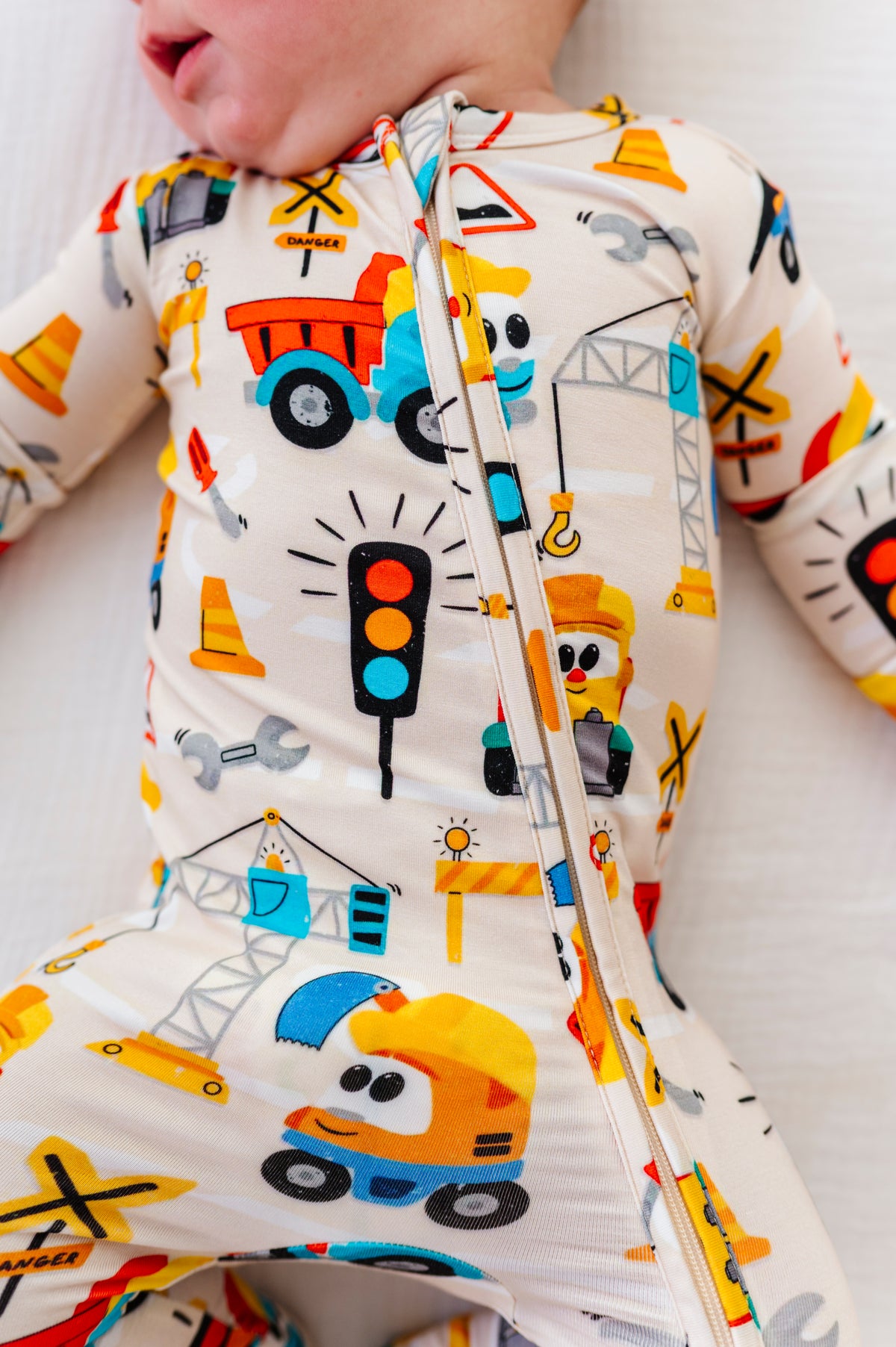 A close-up of a baby wearing a one-piece zip-up sleeper with a colorful construction vehicle print on a cream background.
