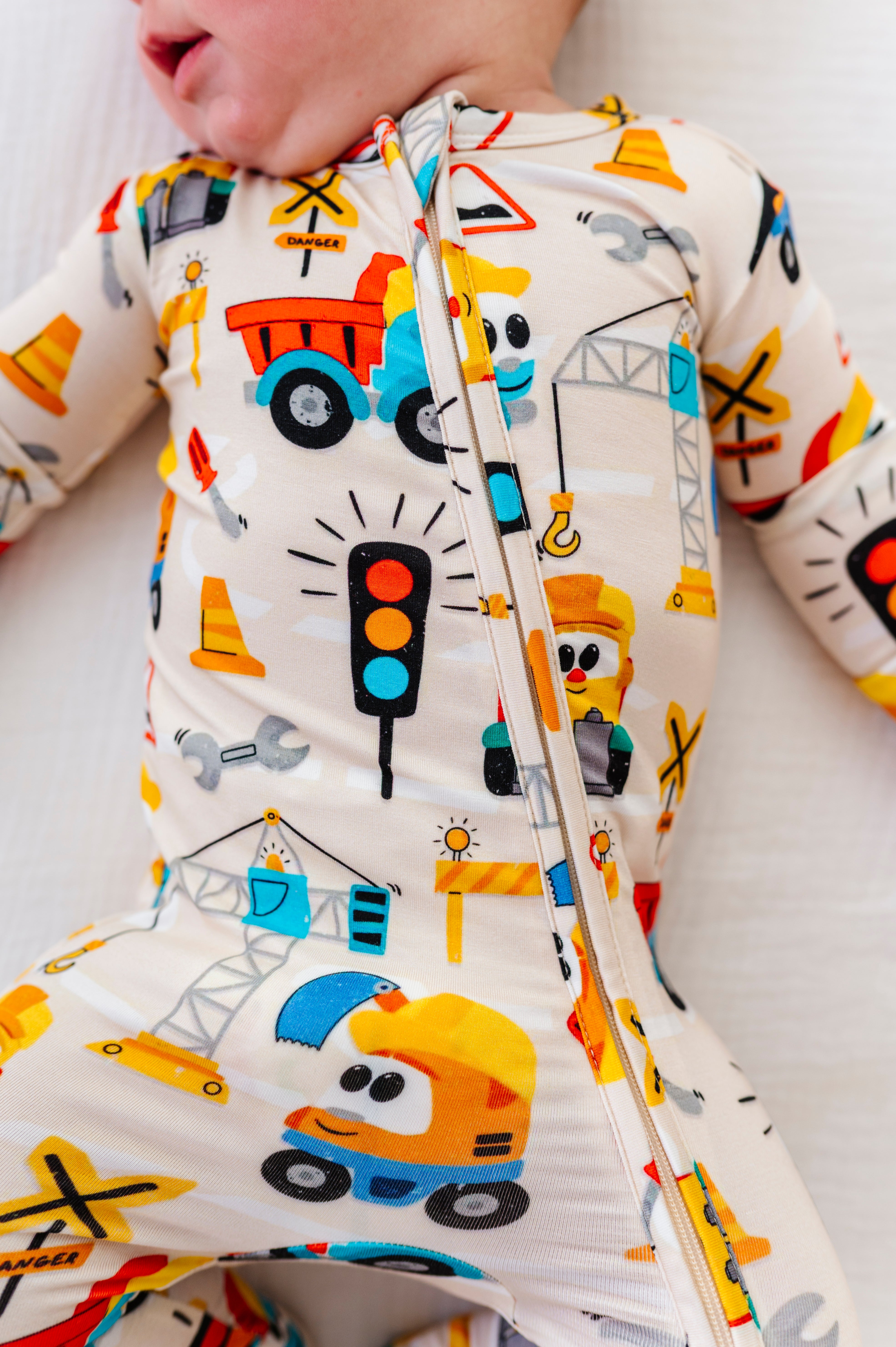 A close-up of a baby wearing a one-piece zip-up sleeper with a colorful construction vehicle print on a cream background.