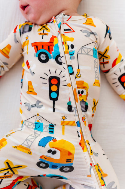 A close-up of a baby wearing a one-piece zip-up sleeper with a colorful construction vehicle print on a cream background.