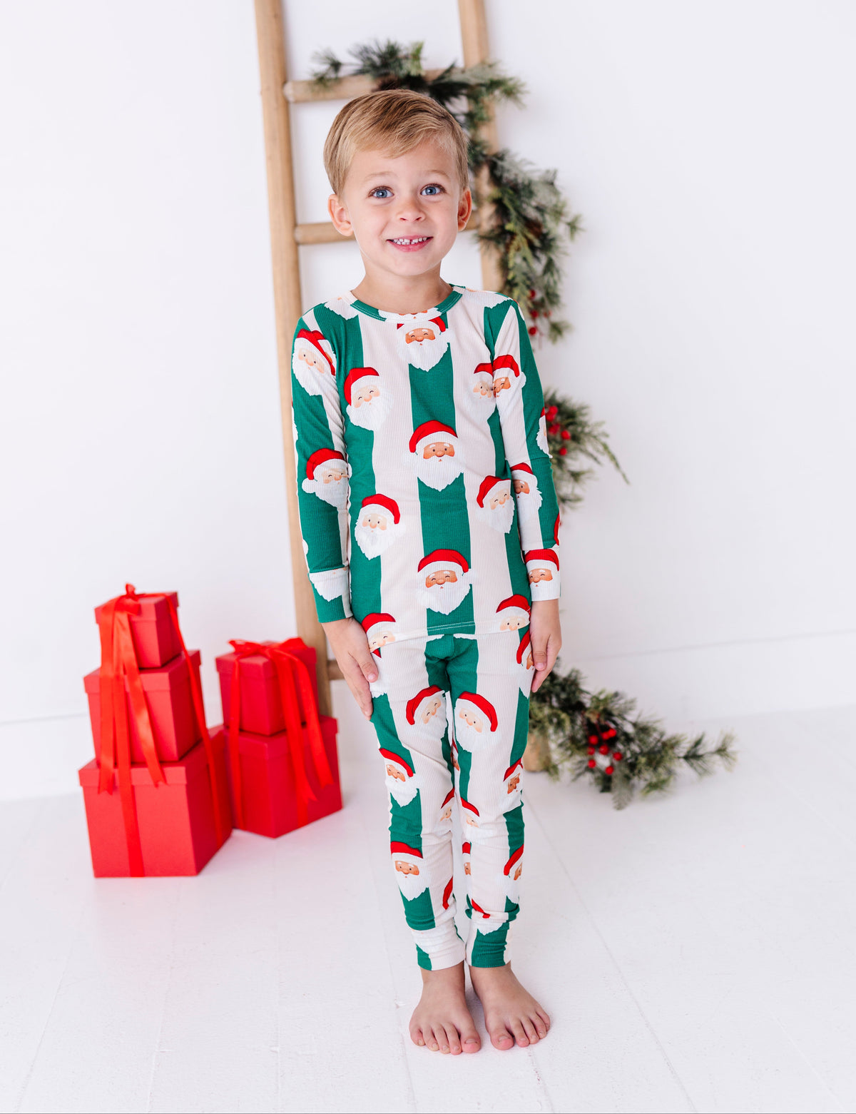 A young blonde boy wearing green-and-beige striped bamboo pajamas with Santa faces all over them. The super soft bamboo pajamas are ribbed and made to fit snugly for safety standards. He's standing in front of Christmas greenery 
