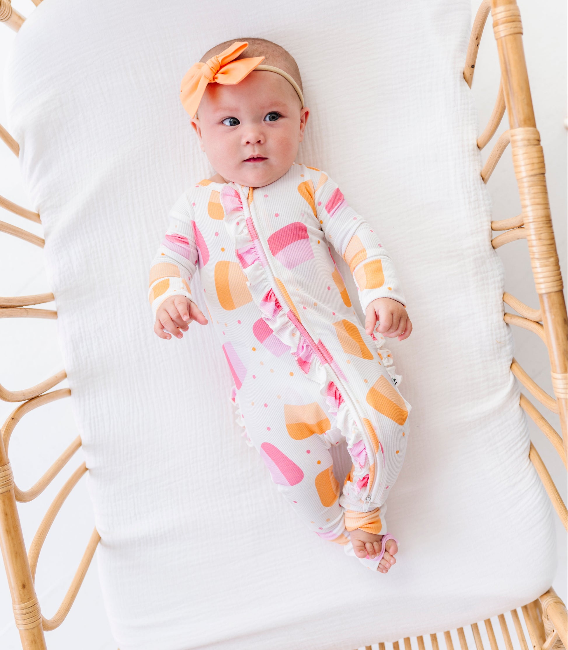 A baby wearing a white footless one-piece sleeper with pink and orange abstract shapes, a matching orange bow headband, and lying on a white cushion inside a rattan bassinet.