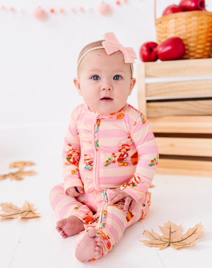 A baby girl sitting on a white floor wearing a pink and white striped footless one-piece pajama with a colorful floral pattern, along with a matching pink bow headband. The background features a wooden crate with red apples and scattered autumn leaves.