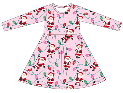 A flat-lay vector illustration showing the front and back views of a long-sleeve children's dress. The dress has a light pink background with a festive Christmas print featuring Santa Claus, reindeer, and glowing holiday lights.