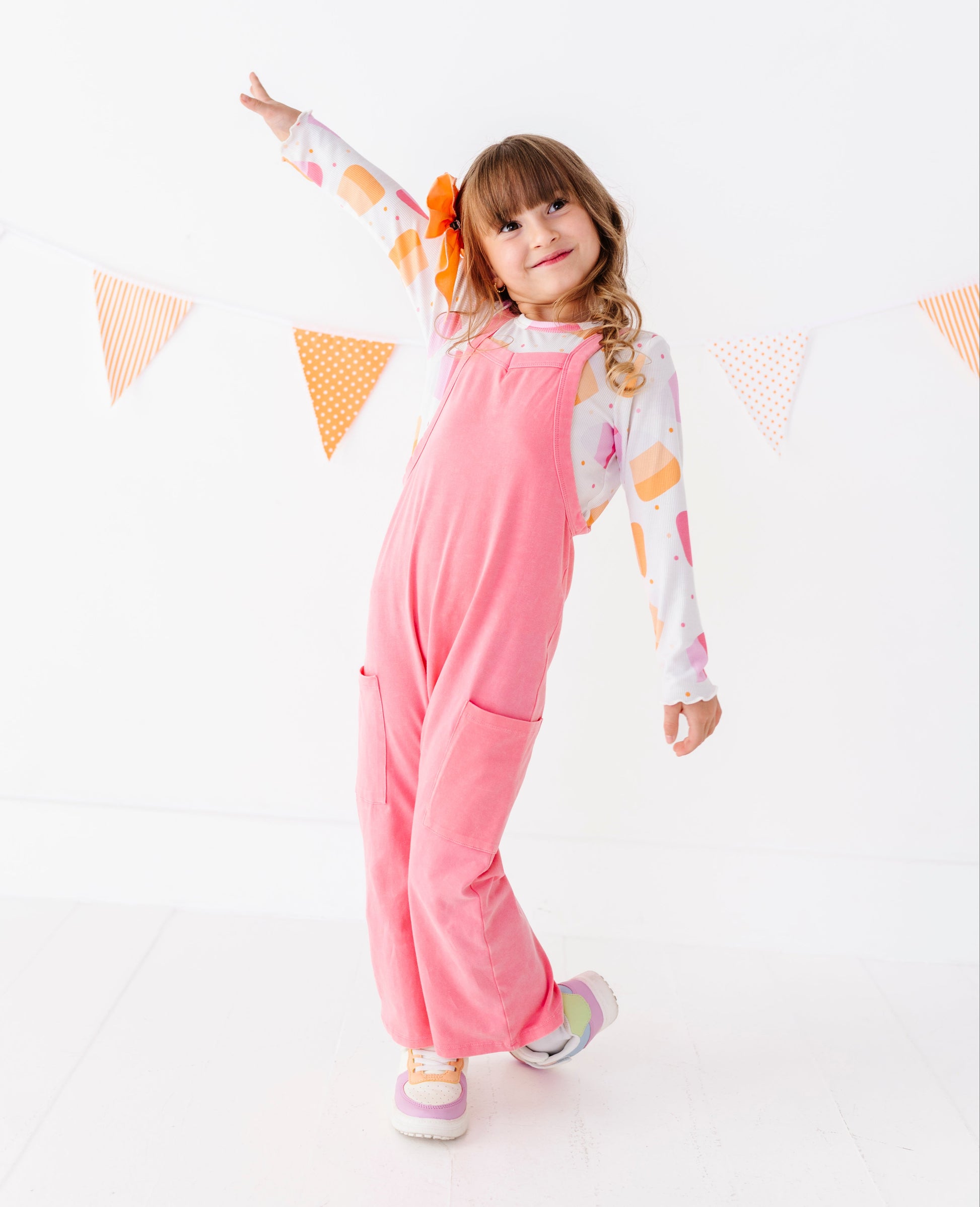 A young girl posing in pink corduroy overalls over a white long-sleeve shirt with colorful illustrations, set against a bright white background with a string of orange and white bunting.