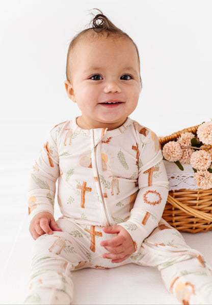Baby wearing a religious patterned romper sitting on a white surface with a basket of flowers in the background.