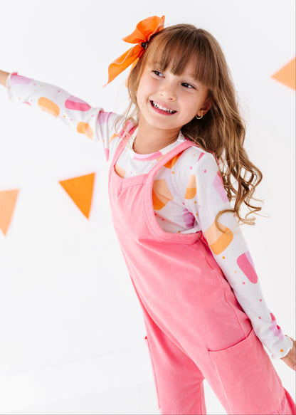 A young girl with a brown hair bow smiles while wearing pink overalls over a white long-sleeve shirt with orange and pink abstract shapes, set against a white background with orange paper flags.