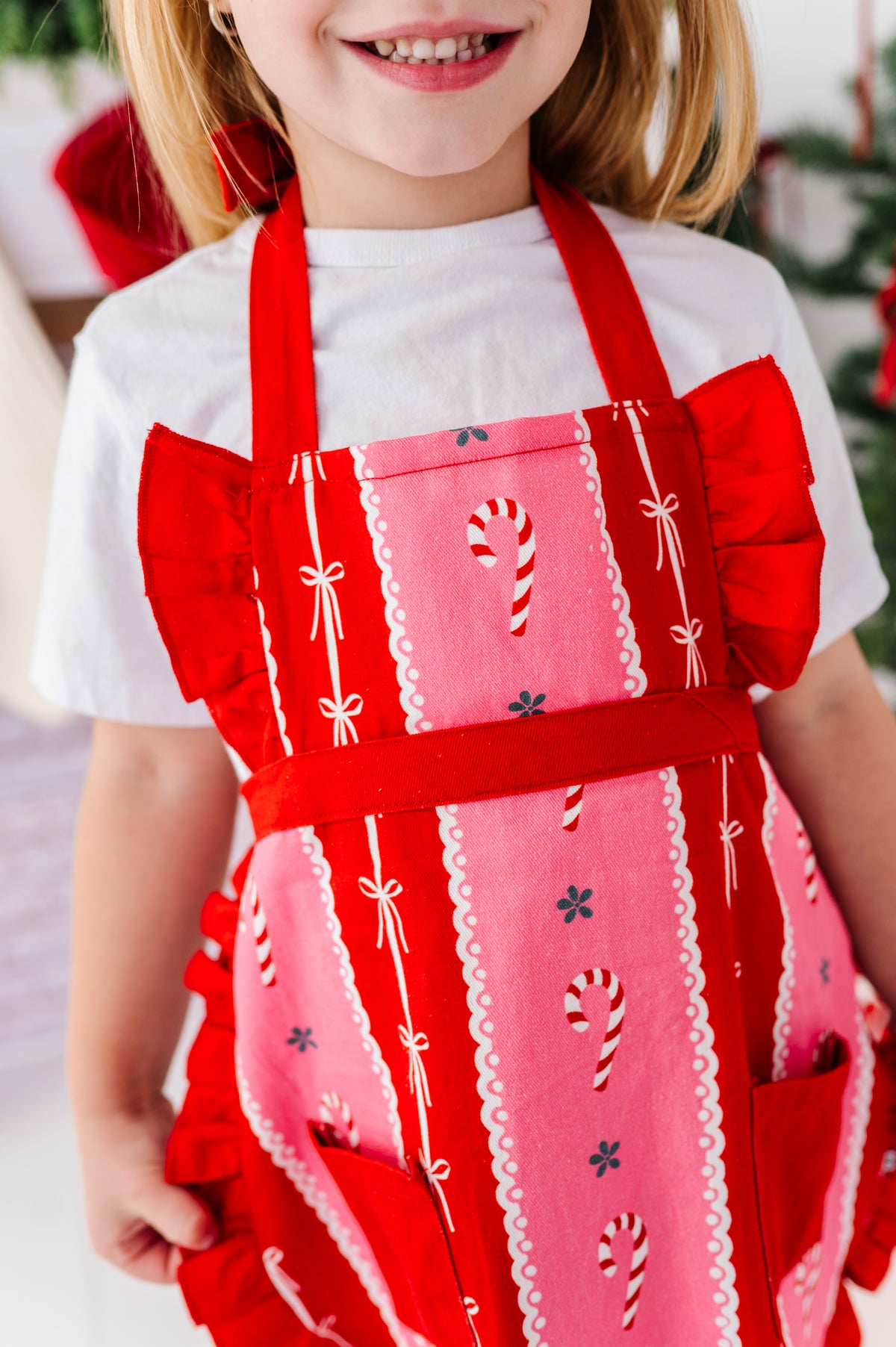 A close-up of a child wearing a festive Christmas apron with red ruffled straps and pockets, and a pink body decorated with candy cane motifs and white lace trim.