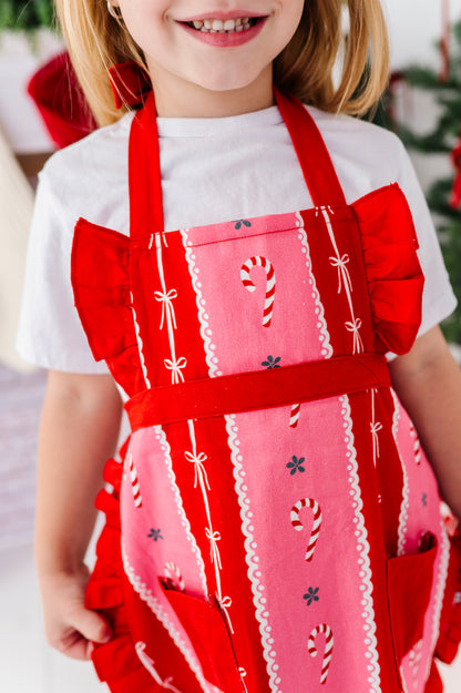 A close-up of a child wearing a festive Christmas apron with red ruffled straps and pockets, and a pink body decorated with candy cane motifs and white lace trim.
