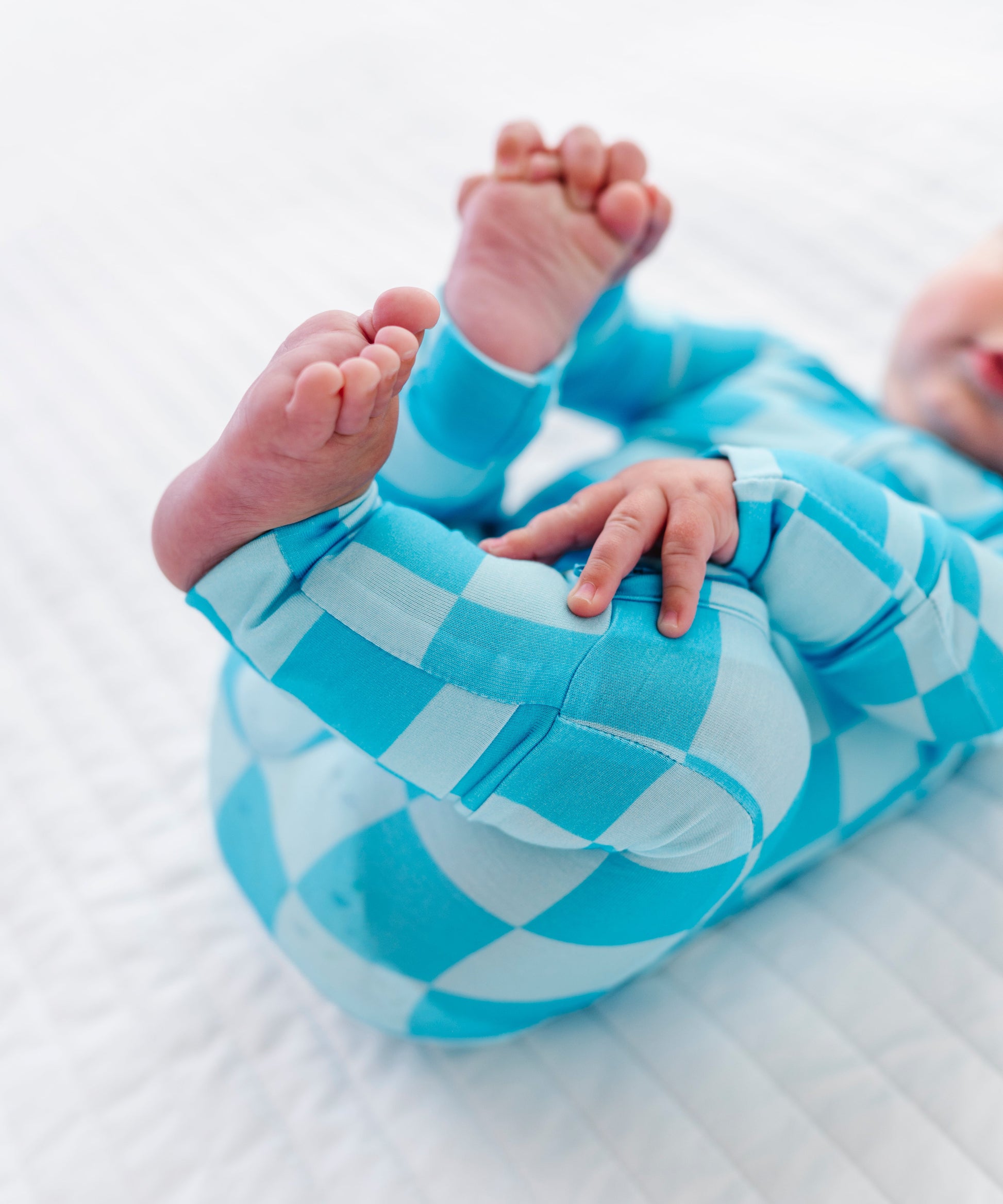A baby lying on a white quilted blanket, wearing a bright blue and white checkered one-piece outfit and kicking their legs in the air.