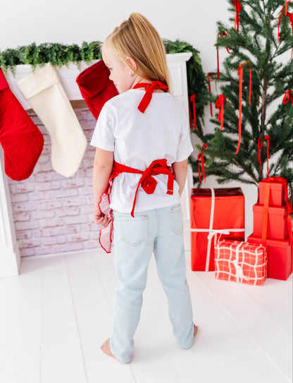 A young girl stands with her back to the camera in a brightly lit holiday room, wearing a white t-shirt with a red bow tied around her waist, light blue jeans, and bare feet. A Christmas tree, stockings, and wrapped gifts are visible in the background.