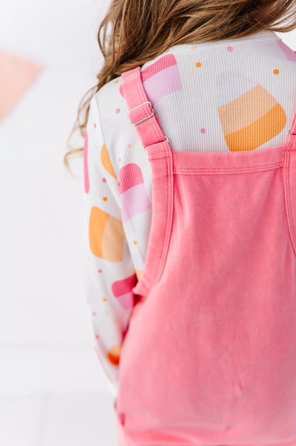 A close-up back view of a child wearing pink sleeveless overalls over a white long-sleeve shirt with a pastel orange and pink geometric pattern.