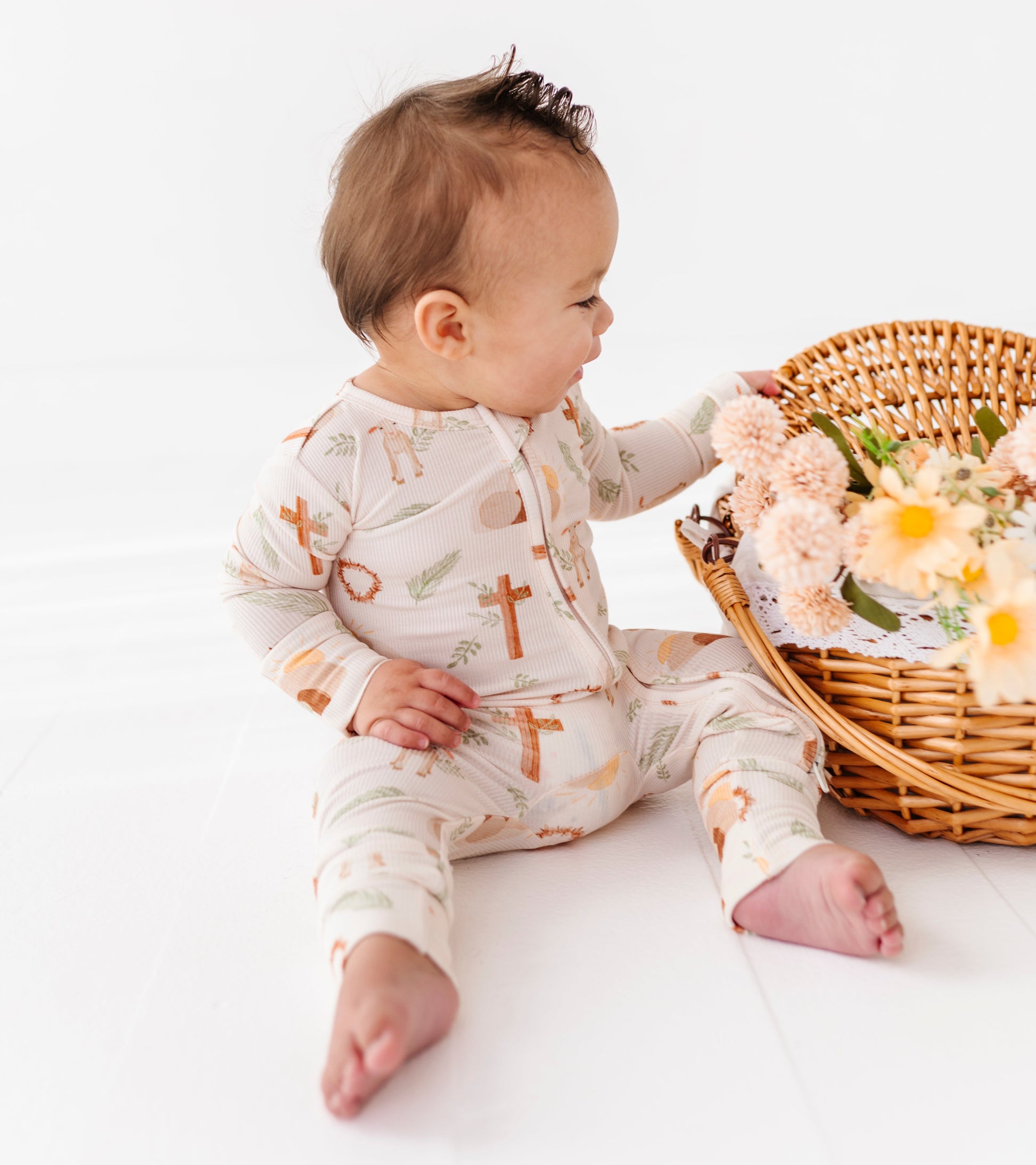 Baby in a patterned Easter romper with brown crosses sitting next to a wicker basket with flowers on a white background