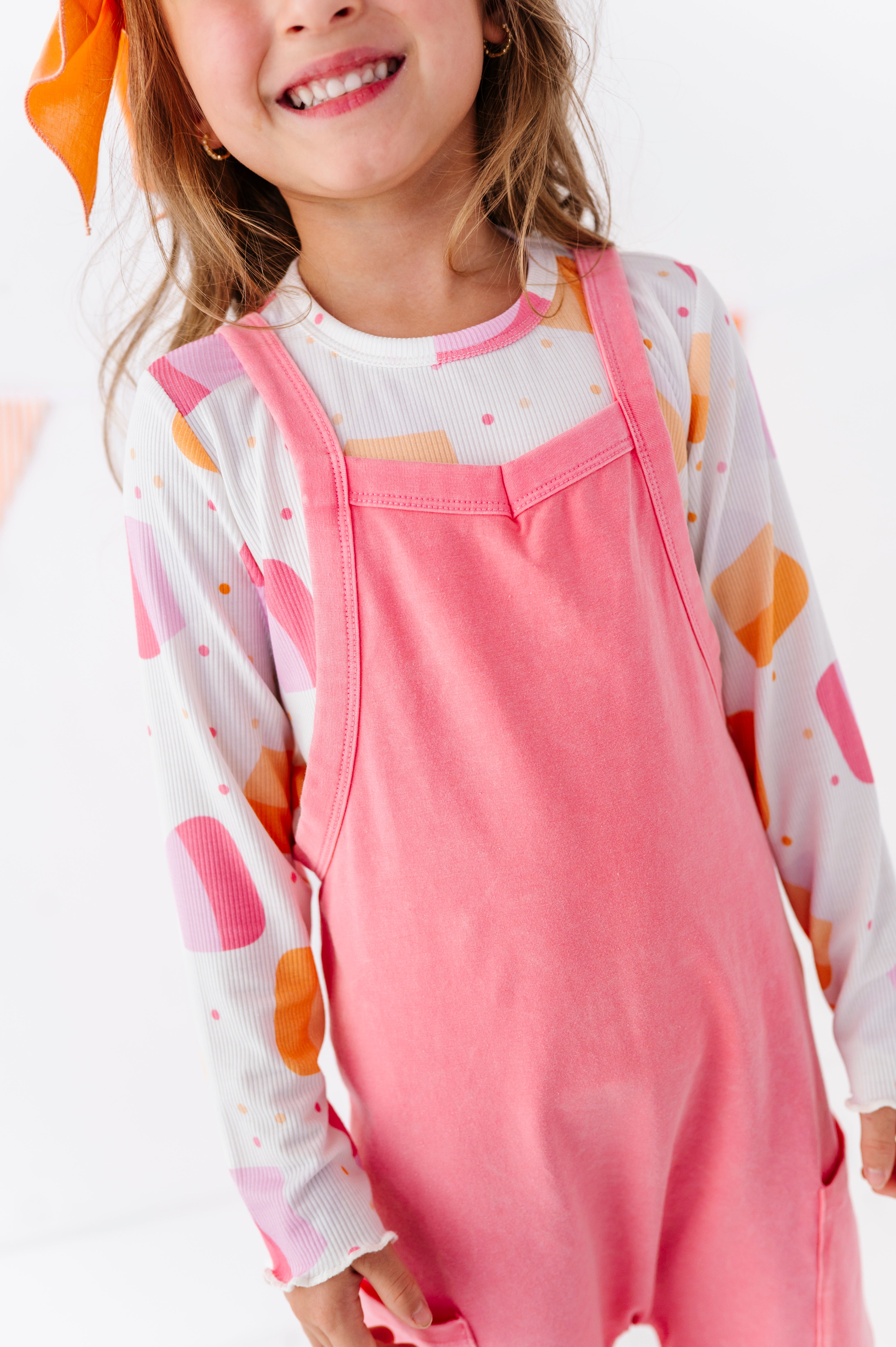 A young girl wearing a pink overall-style jumpsuit over a white long-sleeve shirt with pink and orange abstract shapes, photographed against a clean white background.