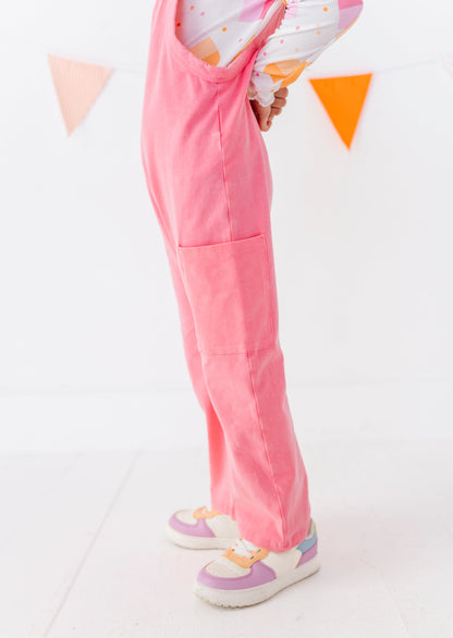 A child wearing bright pink corduroy overall-style pants with a large front patch pocket, shown from the waist down in a studio setting with colorful bunting and pastel sneakers.