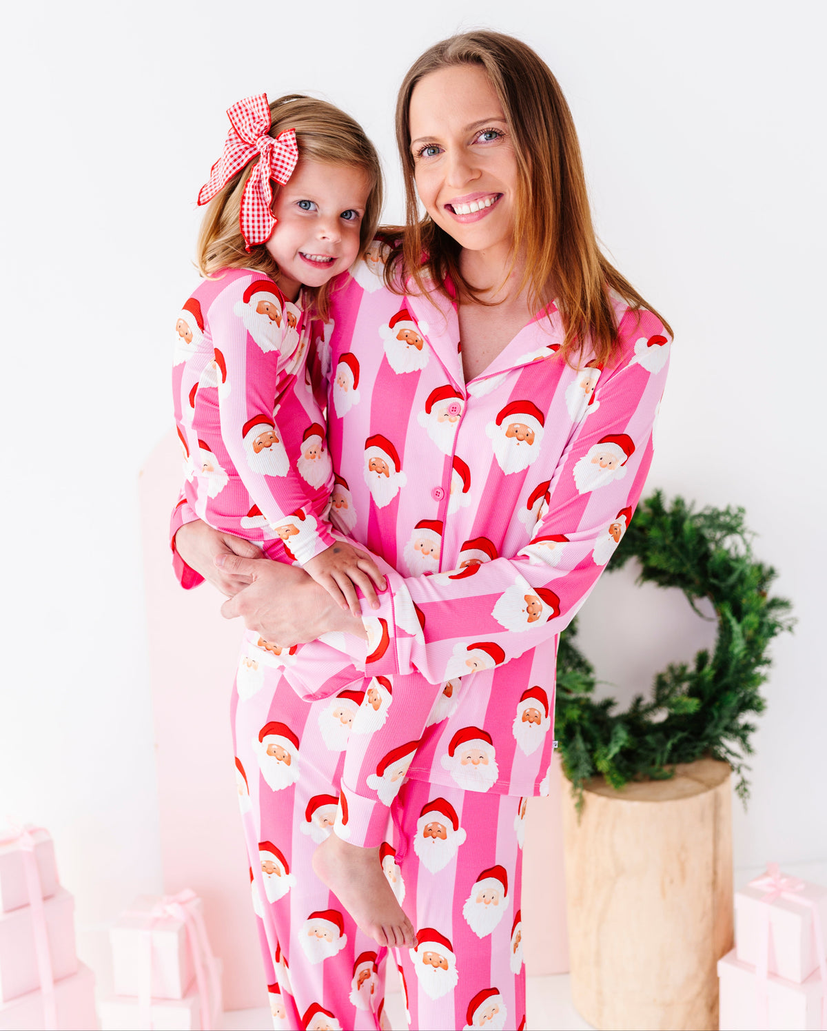 A woman and a young girl wearing matching pink Santa Claus bamboo pajamas, standing together in a bright white room with a green wreath and wrapped gifts in the background.