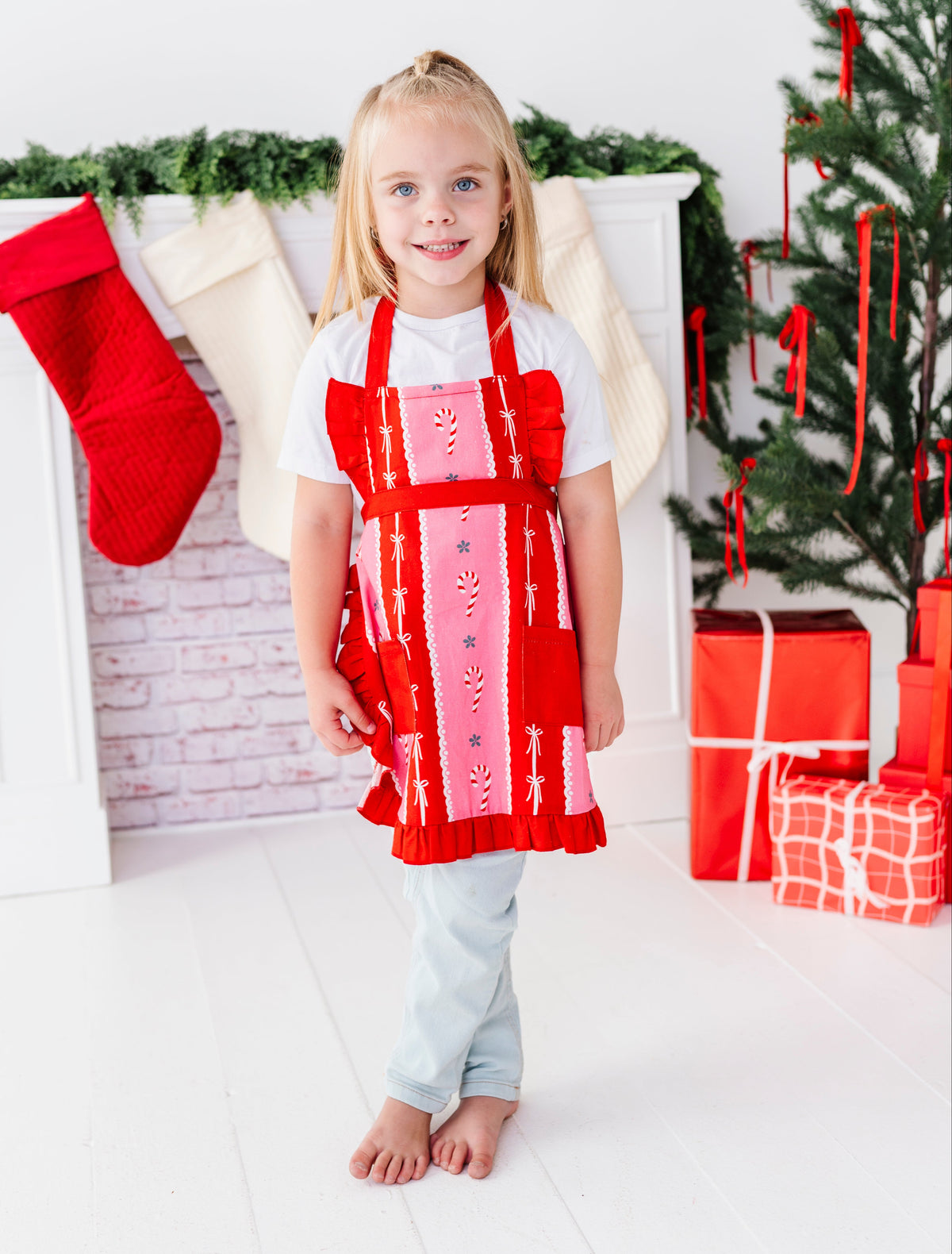 A young girl stands barefoot in a festive holiday setting, wearing a red and pink Christmas apron over a white t-shirt and light blue jeans. The background features a white brick fireplace with a red stocking, green garland, a decorated Christmas tree, and wrapped gifts.